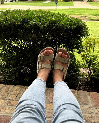 Person wearing beige sandals and light blue jeans standing on a brick path with greenery in the background.